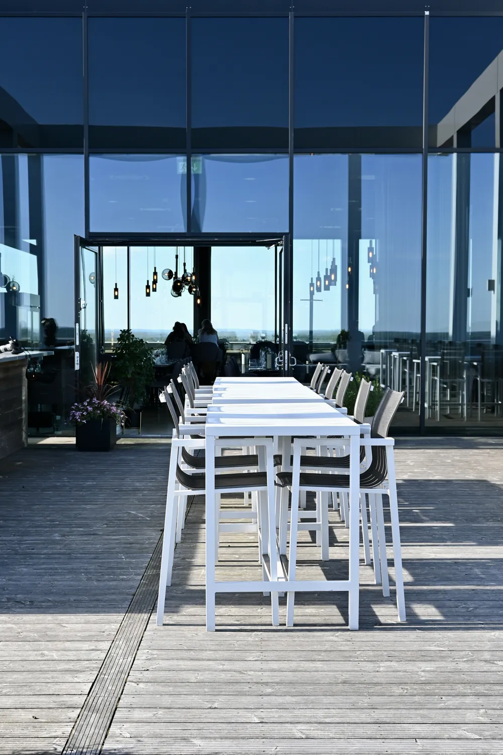 Terrassen på våning 19 med vita barbord framför glasfasaden, dörren öppen mot matsalen och hänglamporna innanför
