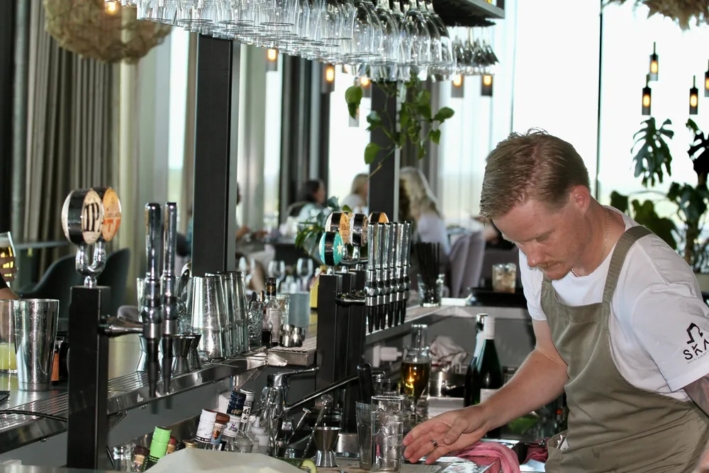 Bartendern bakom disken på SKAI med ölkranarna, glasen i taket och gäster i matsalen bakom